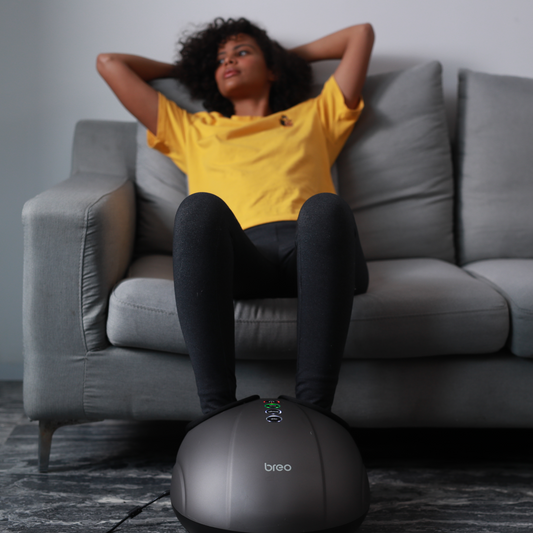 Woman relaxing with Breo foot massager on a gray couch. Focused on foot fatigue relief with the Breo foot massager.